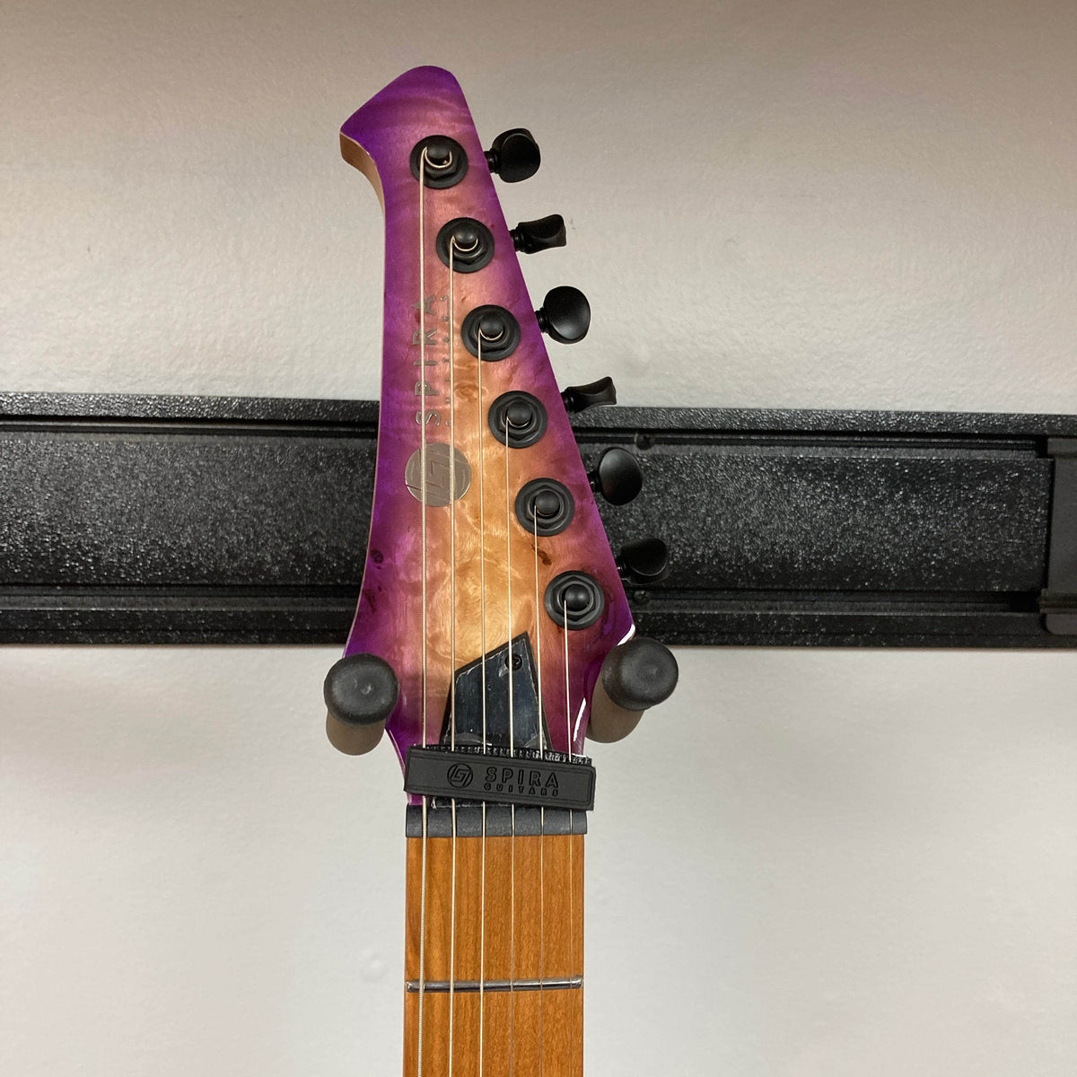 A close-up of the Spira S-450 Trans Purple TPP guitar headstock shows its purple-to-natural wood gradient, six black tuners, a locking nut, and a capo just below the nut. The modern D profile neck by Spira stands out against a white wall background.
