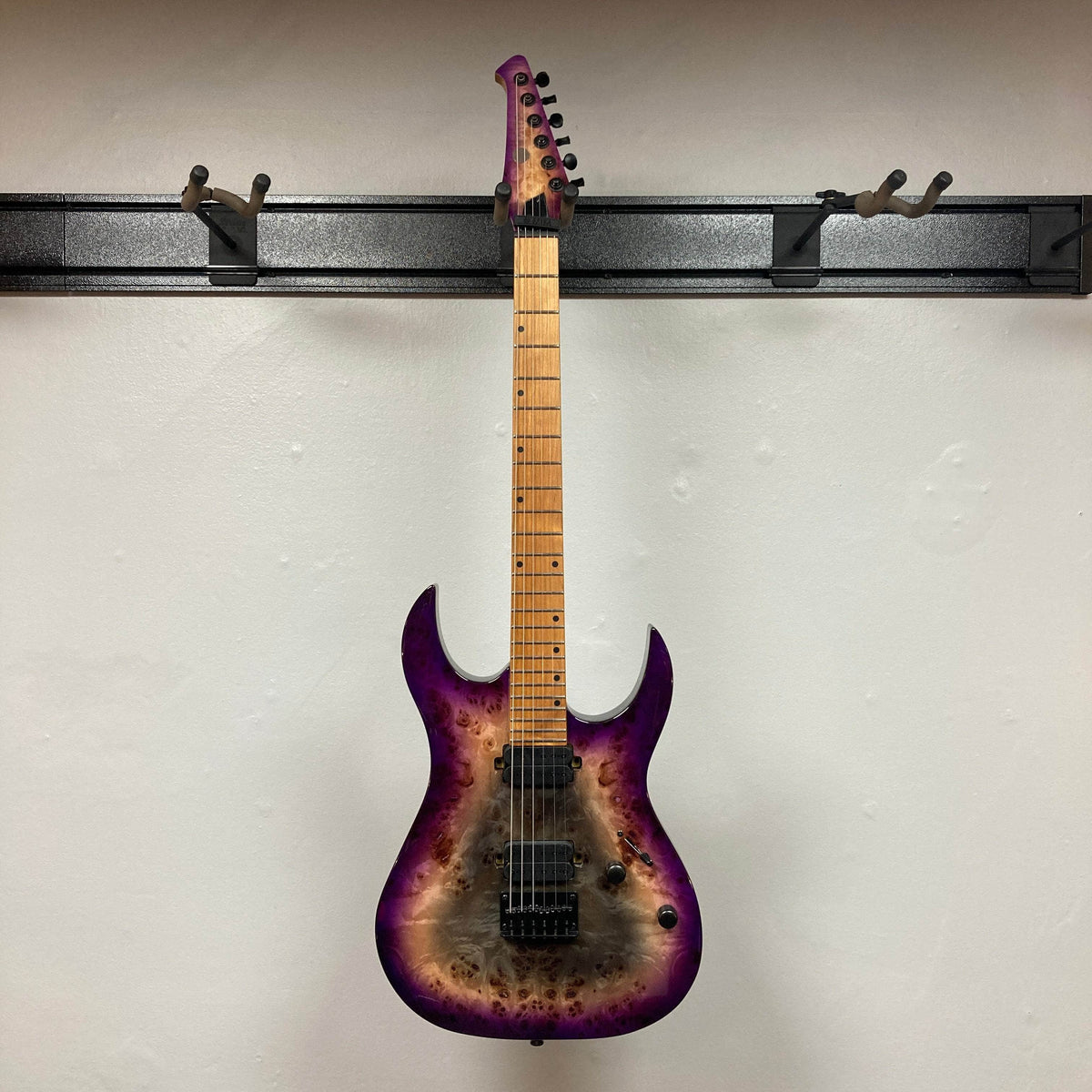 A Spira S-450 Trans Purple TPP seven-string electric guitar by Spira, featuring a maple neck with modern D profile and black hardware, hangs on a wall-mounted rack, its purple and wood burst finish contrasting the white wall.
