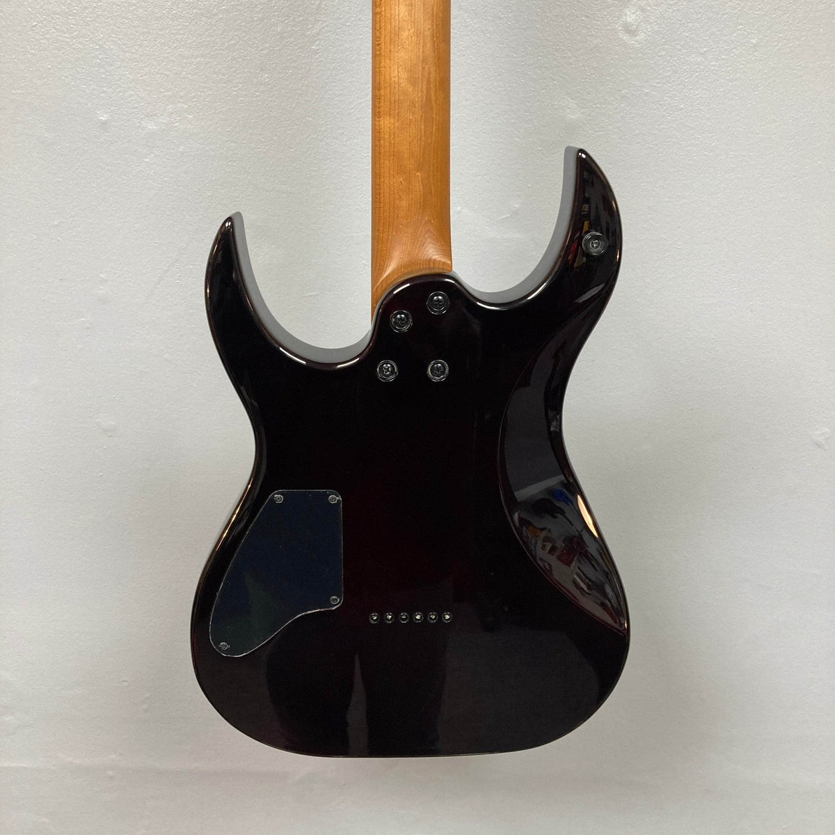 The back of a Spira S-450 Trans Purple TPP electric guitar by Spira shows its dark, glossy finish and modern D profile neck in lighter wood. Several screws and a plastic cover plate are visible, set against a plain white wall.
