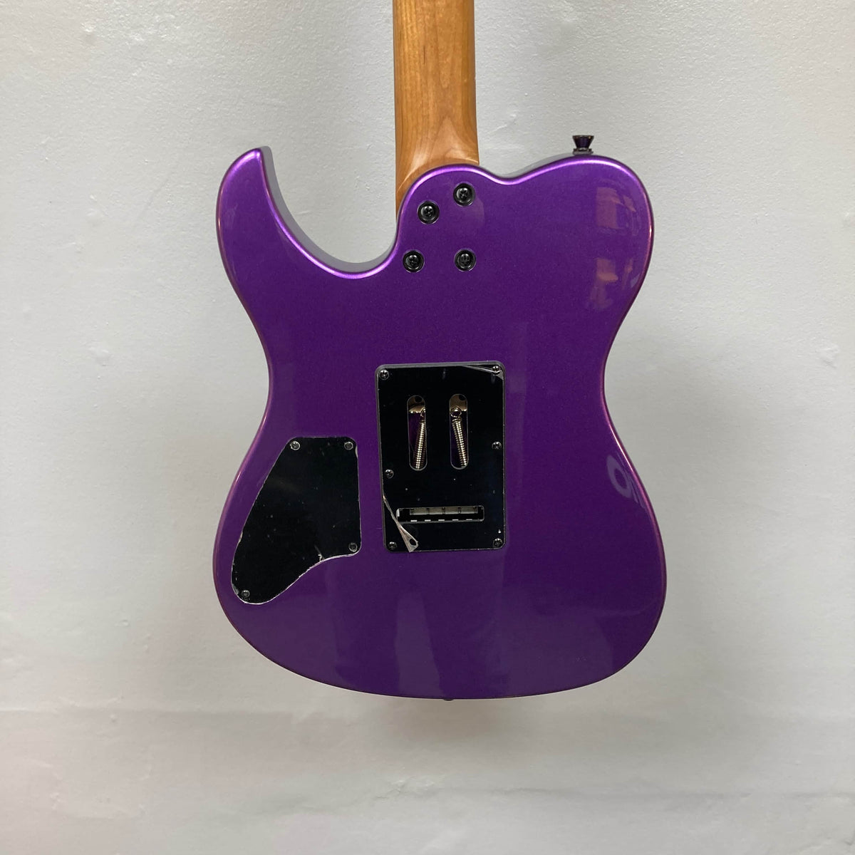The back of a Spira T-420 PP electric guitar by Spira, featuring a purple finish, roasted maple neck, black plastic cover plates, and exposed screws, set against a plain white wall.