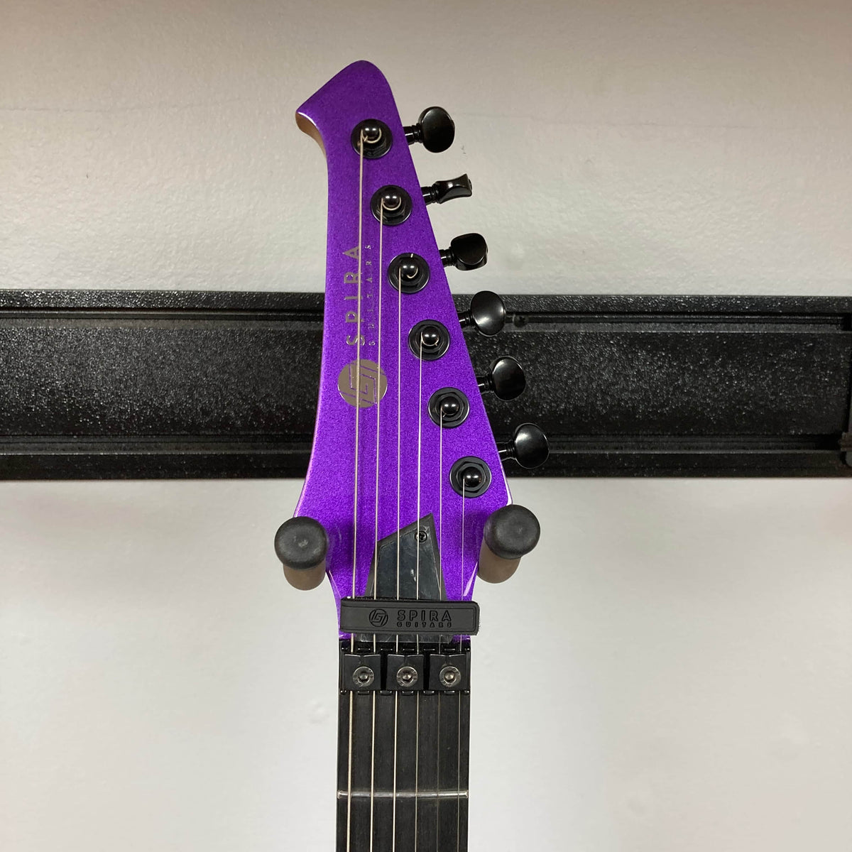 A close-up of a Spira T-420 PP guitar headstock by Spira, in purple with six tuning pegs and a roasted maple neck, mounted on a wall hanger against a white background.