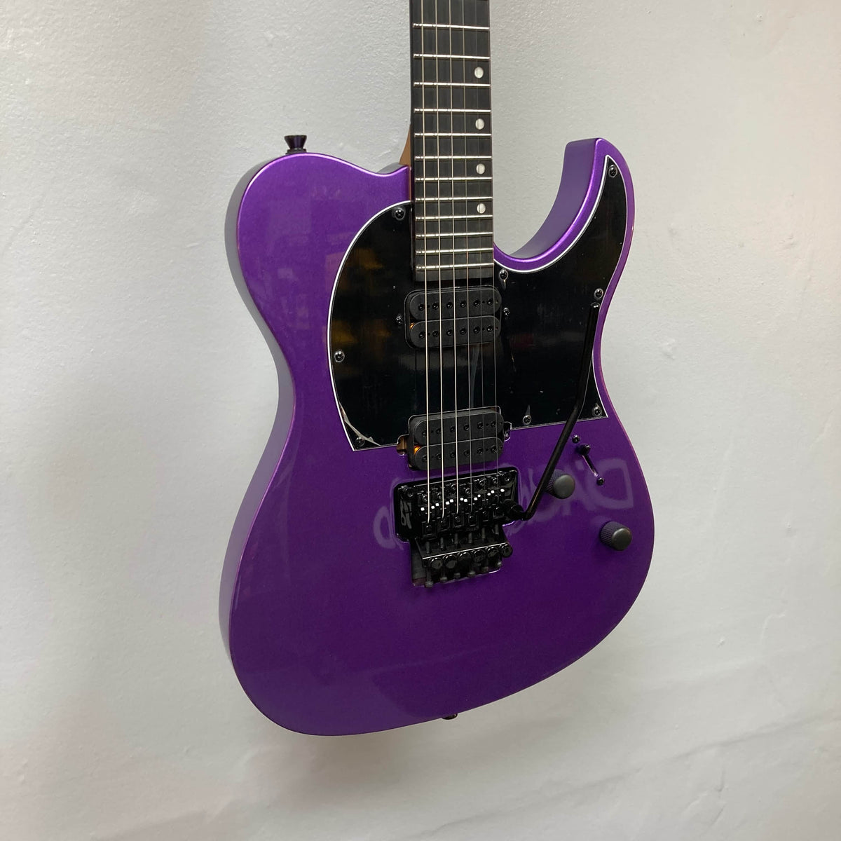 A Spira T-420 PP purple electric guitar by Spira with a roasted maple neck, black pickguard, two control knobs, and a locking tremolo bridge hangs against a white wall.