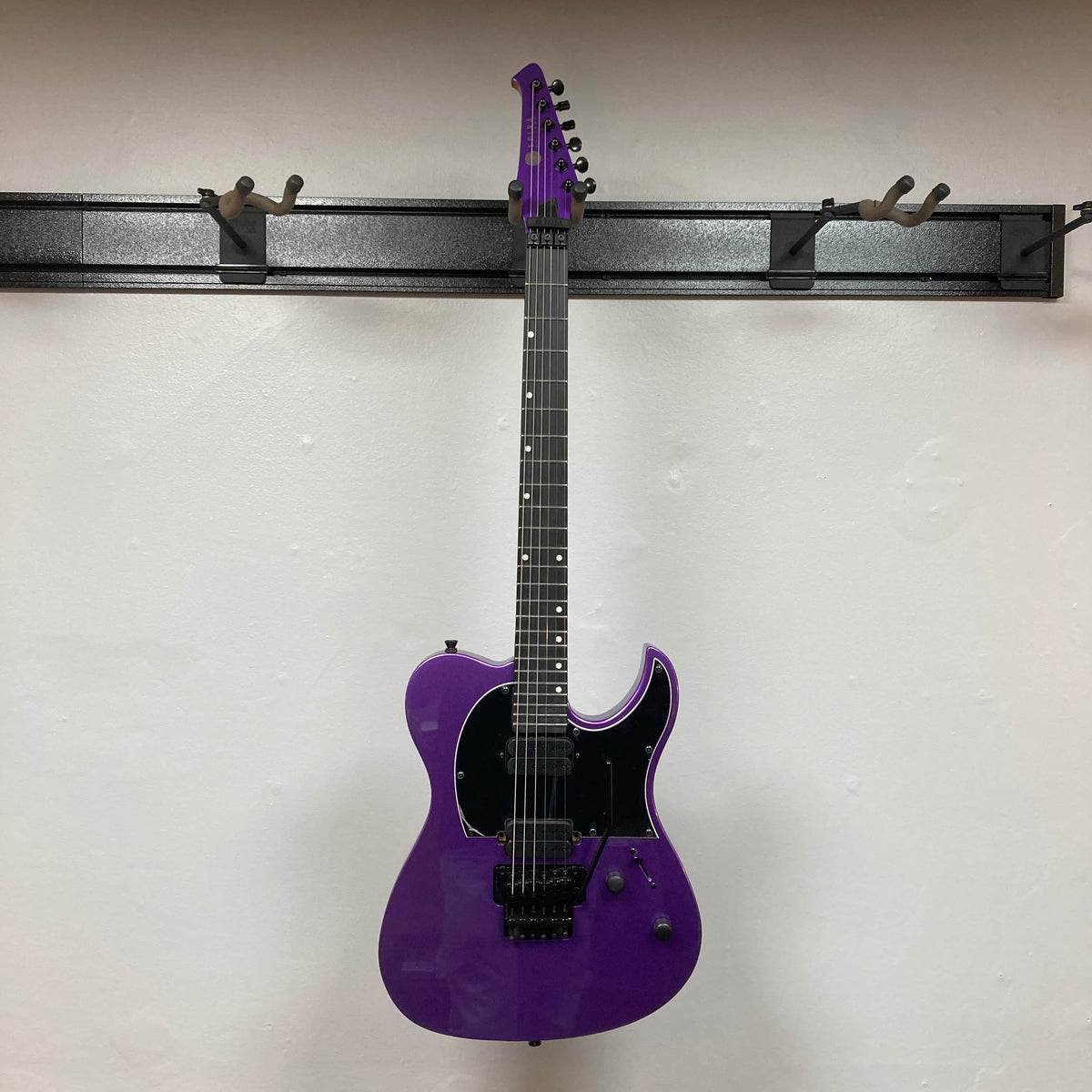 A Spira T-420 PP electric guitar by Spira, featuring a purple finish, black pickguard, six strings, and high-output humbuckers, hangs on a wall-mounted rack against a white wall.