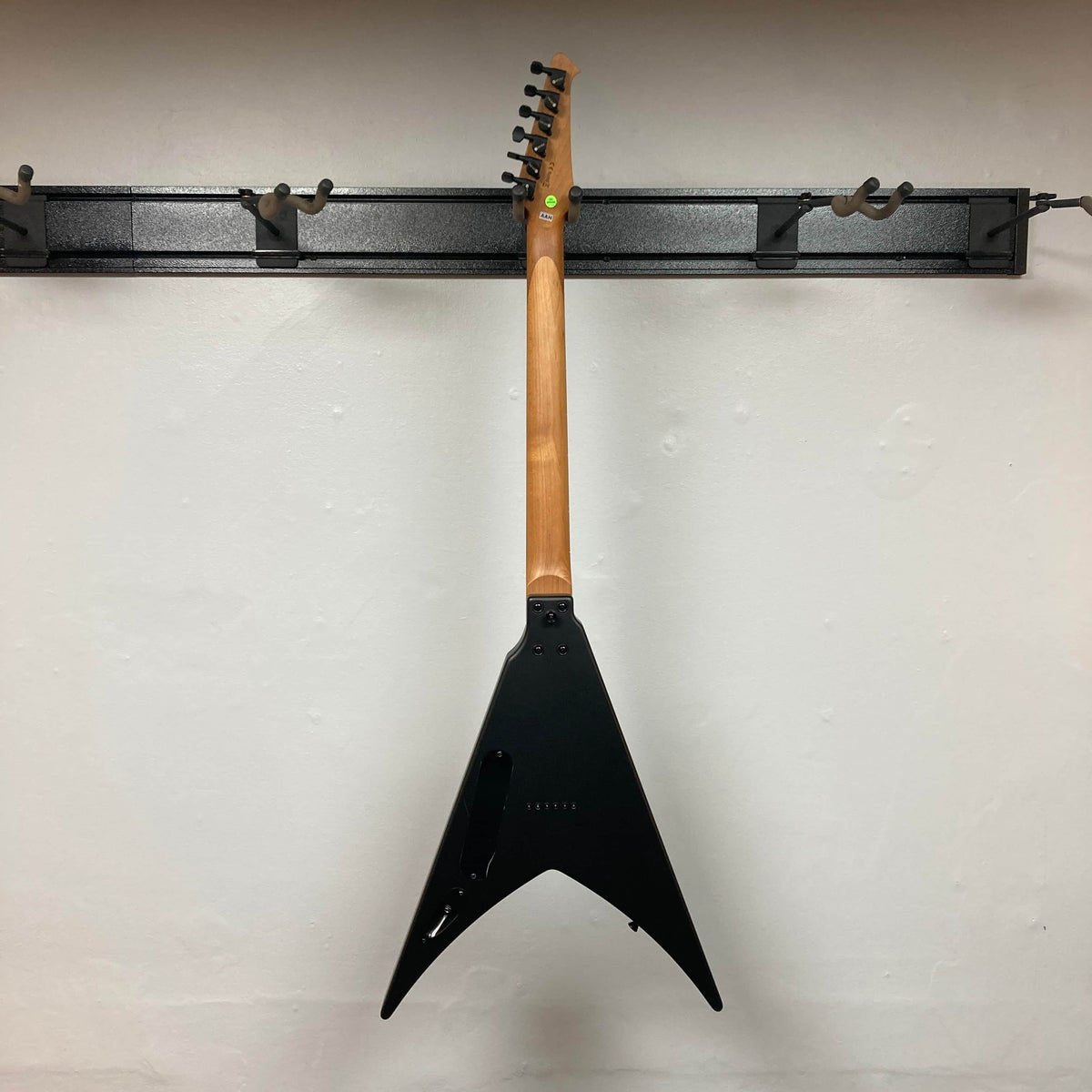A Spira V-400 MBK electric guitar with a black V-shaped body hangs on a wall-mounted rack, shown from the back against a plain white wall—Spira is renowned for tuning stability.