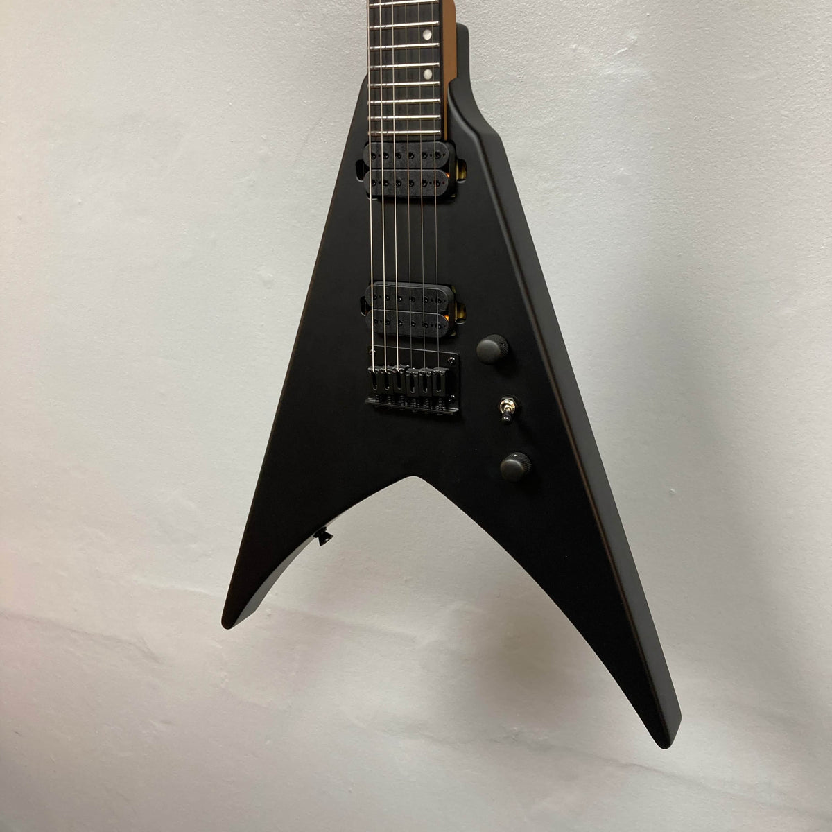 The Spira V-400 MBK electric guitar by Spira features a black V-shaped body, high-output humbuckers, two control knobs, and a toggle switch for stable tuning, displayed against a plain light-colored wall.