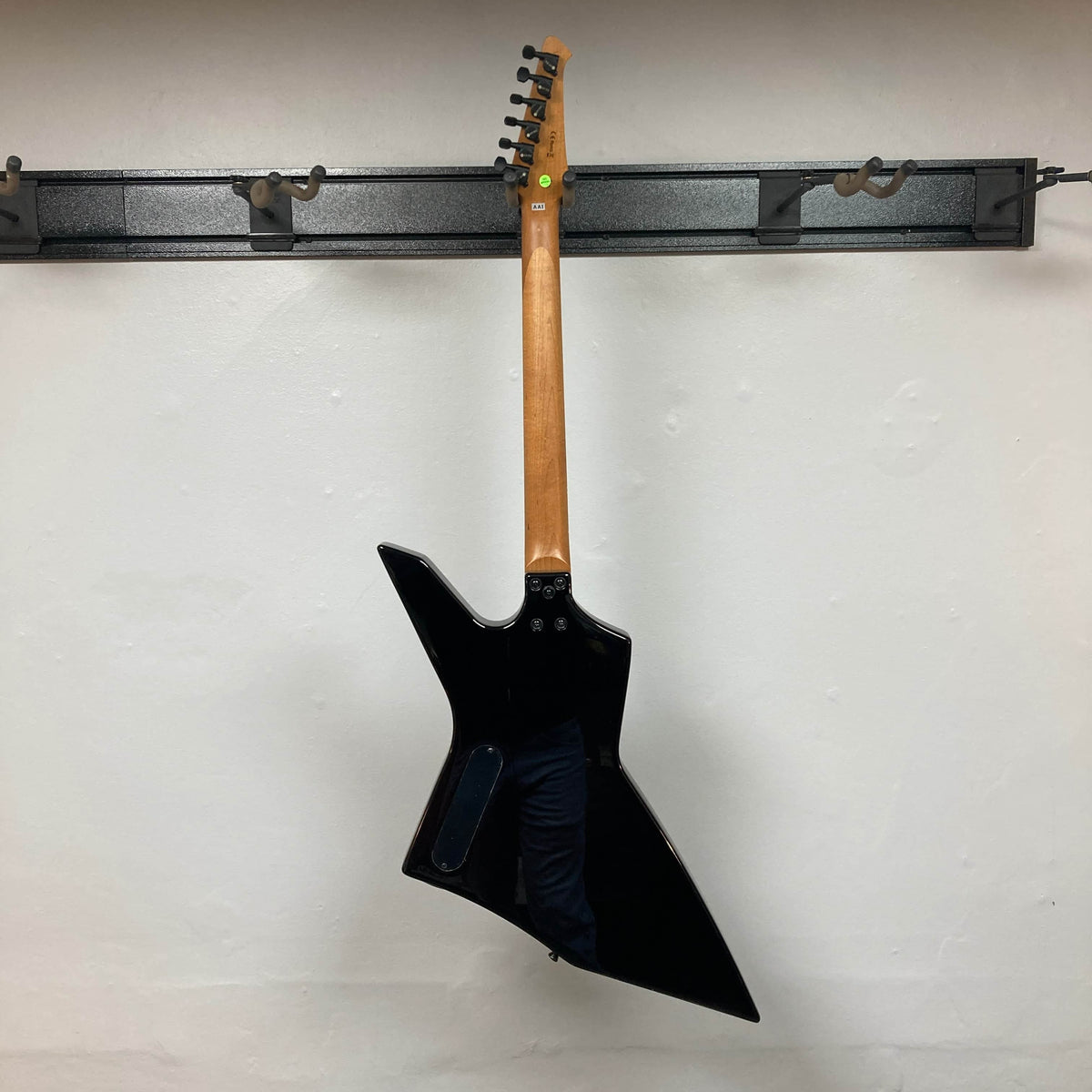 A Spira X-500 TBK E black metal guitar with an angular body and fast neck hangs by its neck on a wall-mounted rack against a plain white wall.