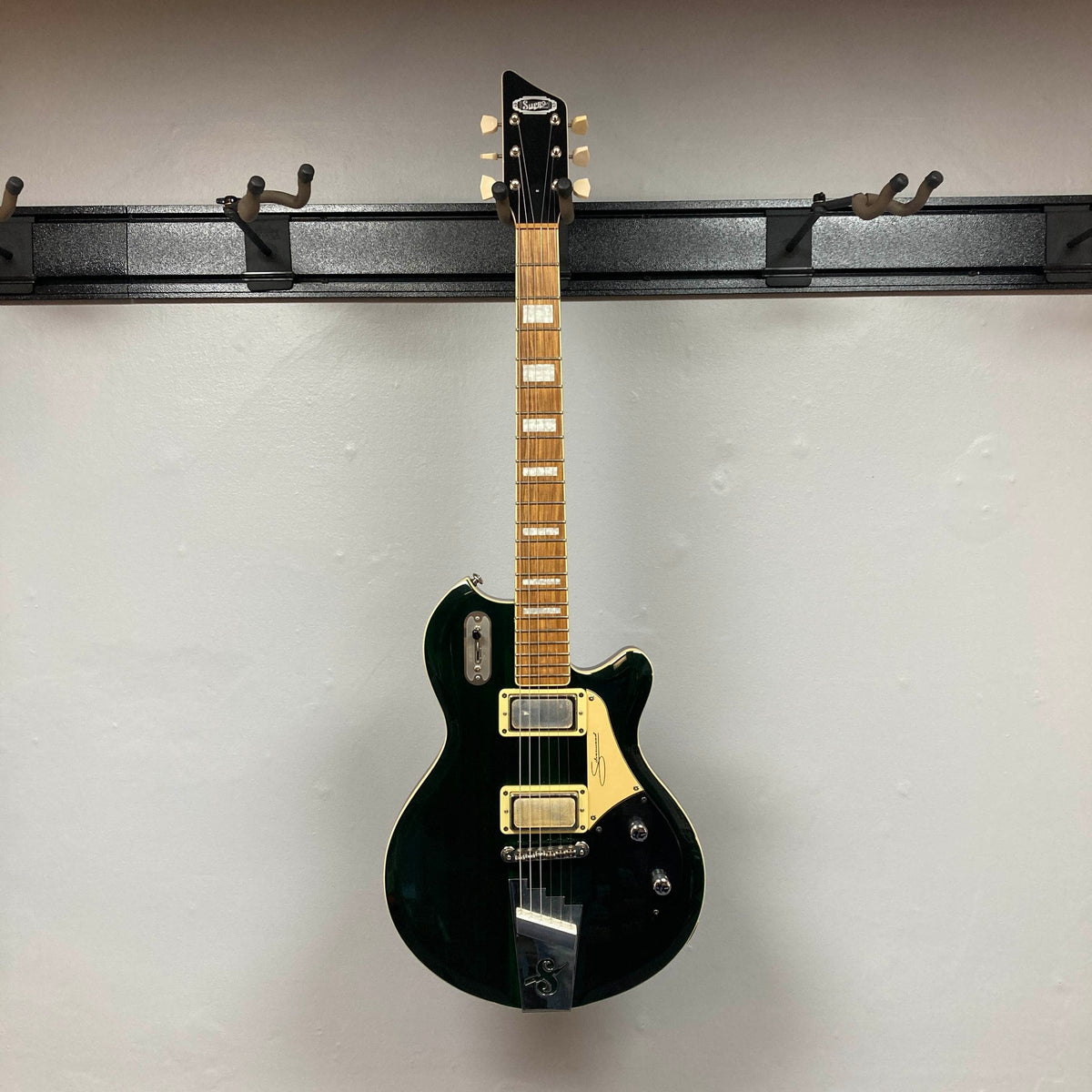 Supro 1296 Silverwood electric guitar in British Racing Green, featuring a dark green body, light wood neck, and unique Gold Foil pickups, is displayed on a wall-mounted black metal rack against a white background. This used Supro guitar blends classic style with modern flair and includes a gigbag.
