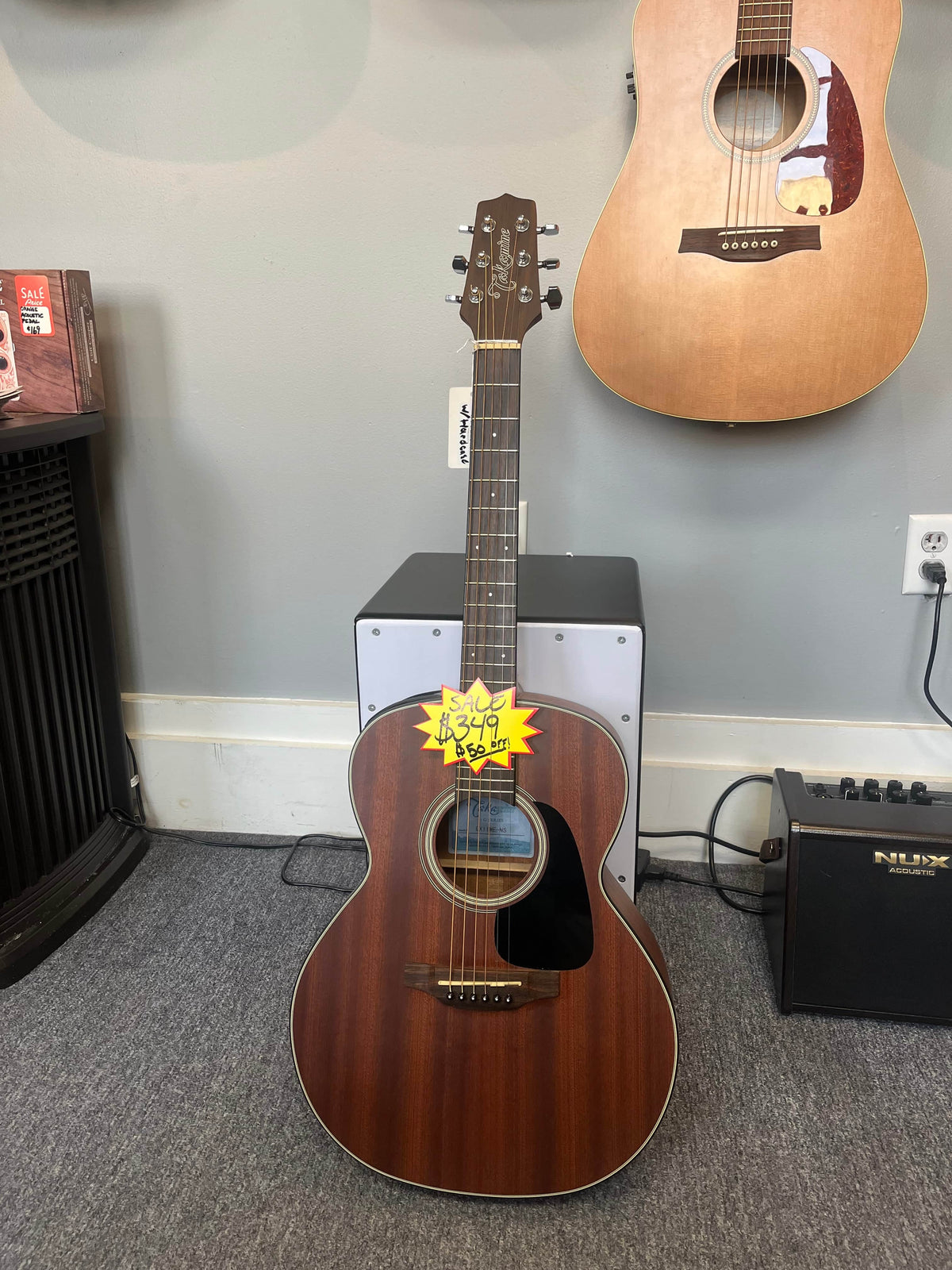 A used Takamine GX11ME Taka-Mini NS 3/4 size acoustic guitar with case is displayed upright on a stand in a music store, with another guitar hanging on the wall and an amplifier nearby.