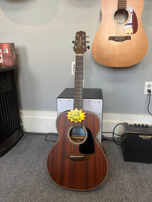A used Takamine GX11ME Taka-Mini NS 3/4 size acoustic guitar with case is displayed upright on a stand in a music store, with another guitar hanging on the wall and an amplifier nearby.