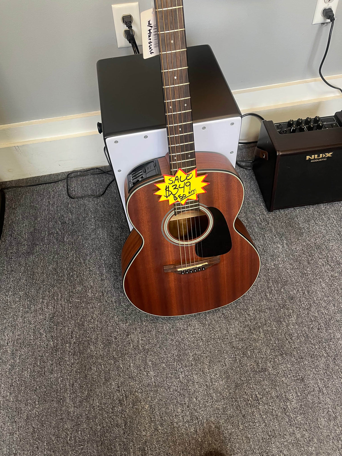 A Takamine GX11ME Taka-Mini NS 3/4 Size Acoustic Guitar w/Case USED/consign is displayed on a gray carpet with a yellow sale tag marked $299, alongside an amp and small black stool—ideal for portable music anywhere.