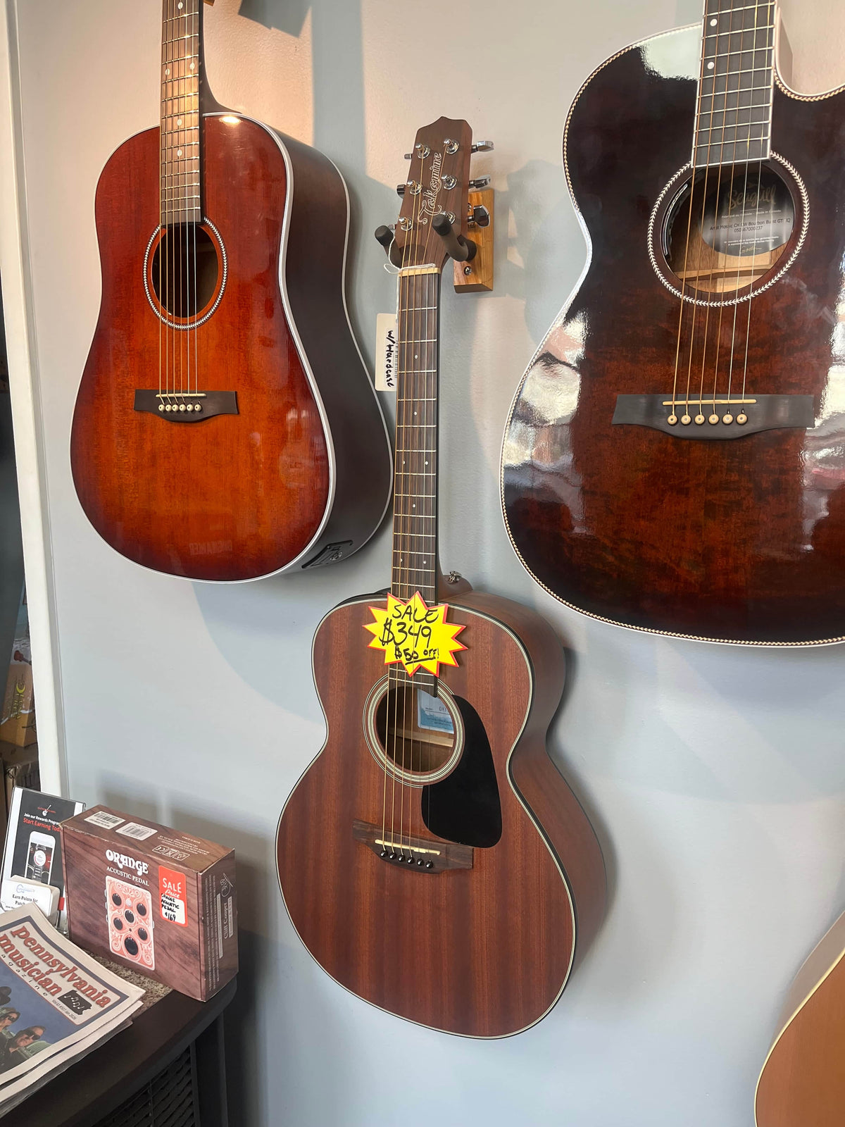 Three acoustic guitars hang on a store wall; the center one is a Takamine GX11ME Taka-Mini NS 3/4 Size Acoustic Guitar w/Case (USED/consign) with a yellow $349 sale tag. A magazine and pedal box below emphasize this portable Takamine’s appeal.