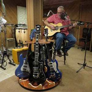A man plays an acoustic guitar and sings in a studio, with several electric guitars displayed on the A&S Crafted Products Carousel™ Deluxe Rotating Guitar Stand | Combined Unit in the foreground.