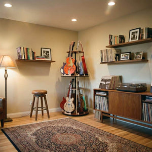 A cozy music room includes the A&S Crafted Products The Carousel™ Deluxe Rotating Guitar Stand | Expansion Tier for efficient guitar storage, shelves with books and records, a turntable with vinyl storage, a stool, floor lamp, and a patterned rug.