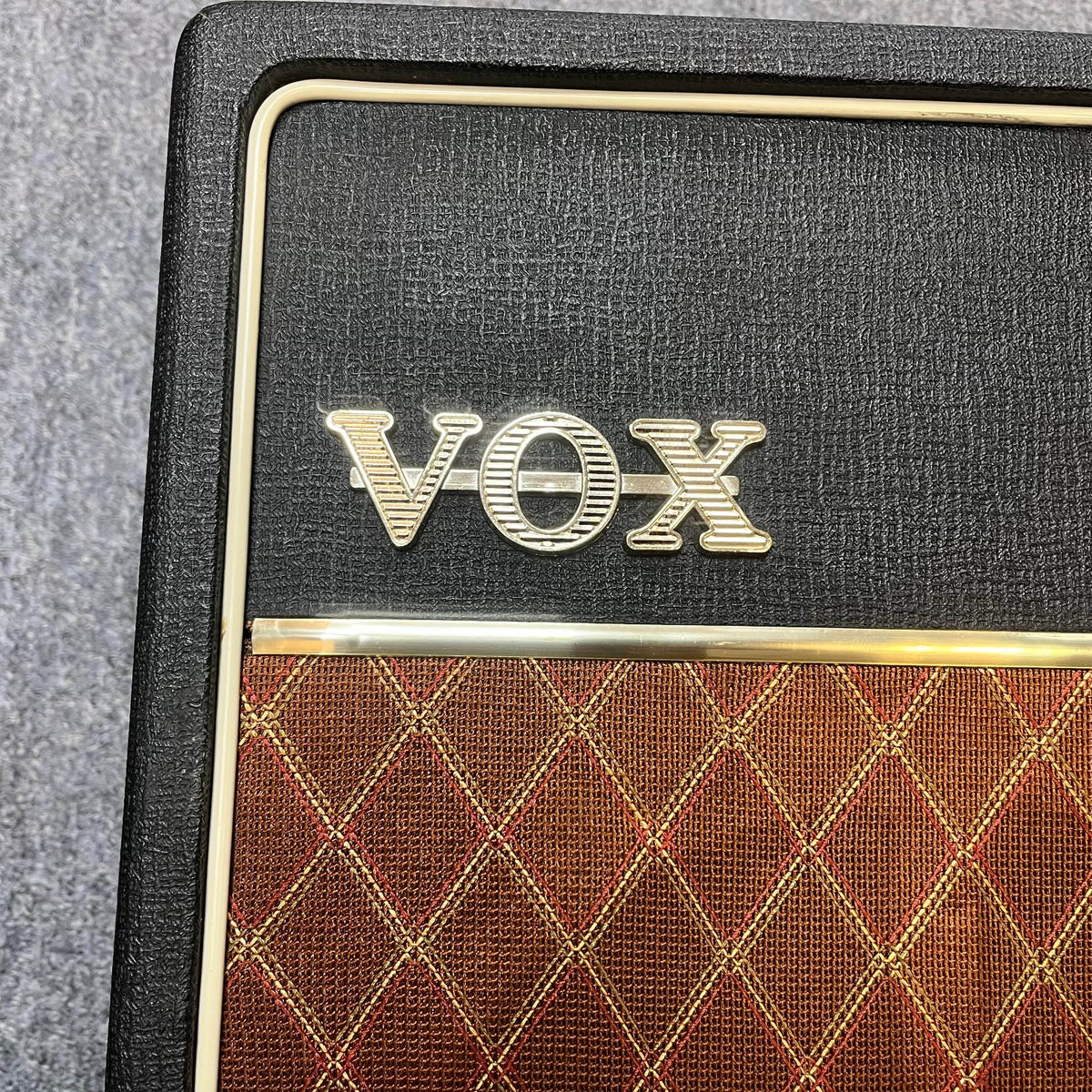 Close-up of a Vintage 1963 Vox AC30/6 Normal amplifier with the Vox logo on textured black above a brown and gold diamond-patterned grille, including its original JMI egg footswitch and flight case.