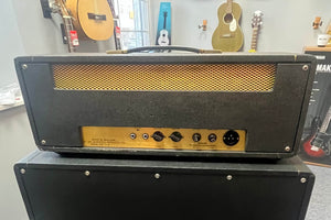 A 1971 Marshall Lead Head JMP 1987 50 Watt amp head sits on a matching cabinet. Multiple control knobs and gold mesh highlight its vintage style, with guitars behind it, capturing classic British grind. Flight case included.