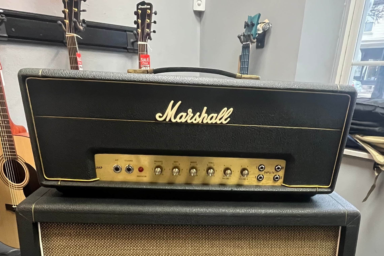 The Marshall 1971 50W Lead Head, JMP Era, with Flight Case rests on a speaker cabinet, showcasing its iconic British sound through its controls. Marshall guitars are displayed in the background. The Marshall 1971 50W Lead Head, JMP Era, with Flight Case rests on a speaker cabinet, showcasing its iconic British sound through its controls. Marshall guitars are displayed in the background.