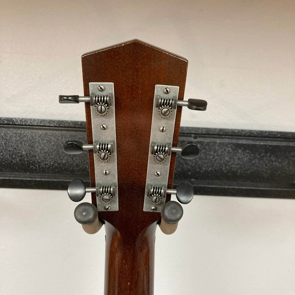 The back of a Waterloo WL-12 Parlor Acoustic guitar headstock, featuring six metal tuning pegs and a dark brown wood finish, is shown against a white wall and black rack. Brand: Waterloo. Model: WL-12 Parlor Acoustic w/Case Used Consign.