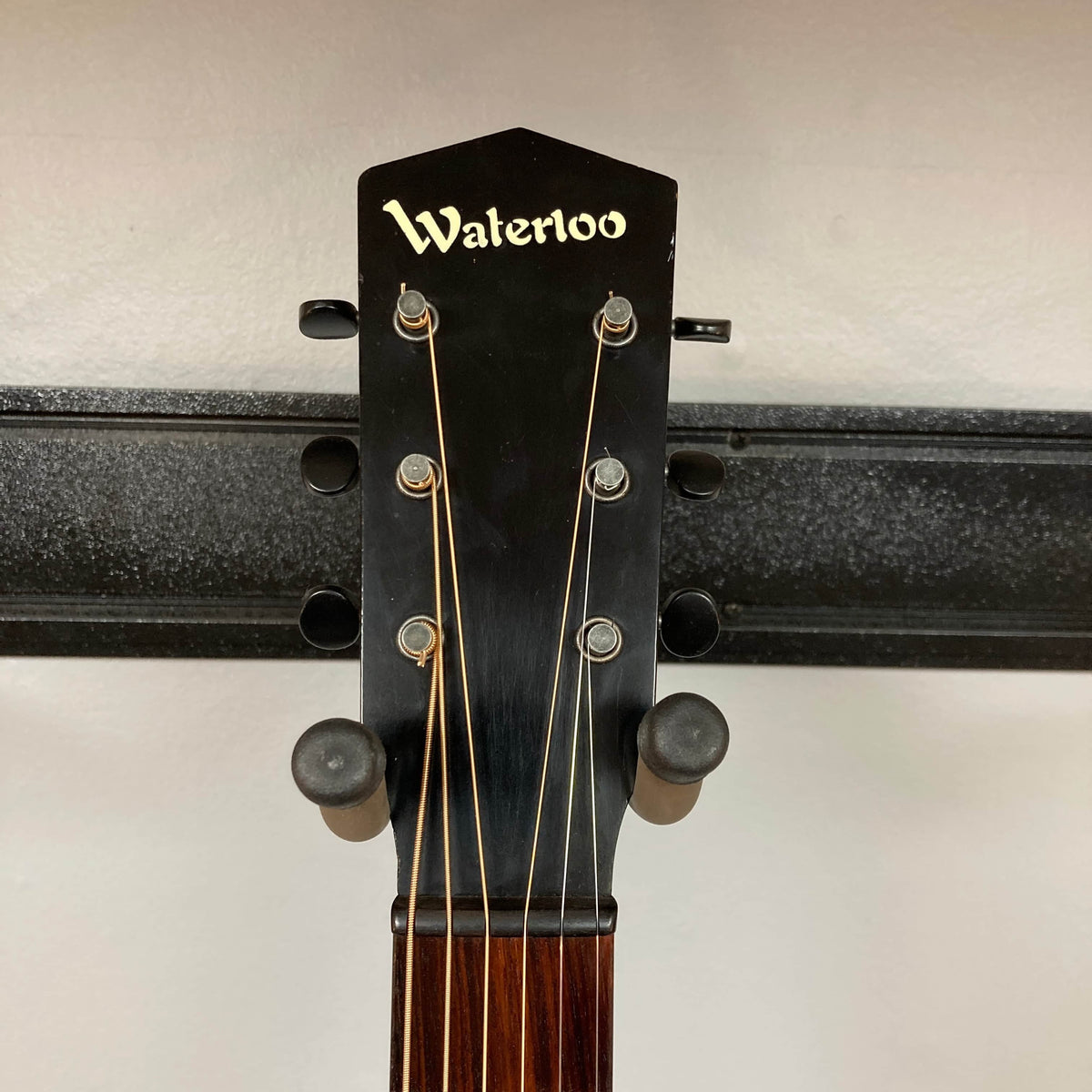 Close-up of the Waterloo WL-12 Parlor Acoustic w/Case Used Consign guitar headstock, featuring "Waterloo" branding, six tuning pegs, and strings, mounted on a wall rack against a light-colored background.