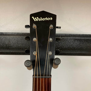 Close-up of the Waterloo WL-12 Parlor Acoustic w/Case Used Consign guitar headstock, featuring "Waterloo" branding, six tuning pegs, and strings, mounted on a wall rack against a light-colored background.