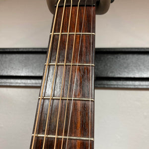 Close-up of a Waterloo WL-12 Parlor Acoustic guitar neck showing six strings, wooden fretboard with metal frets, and part of a capo. A black wall hanger and light-colored wall are visible in the background. Used Consign w/ Case.