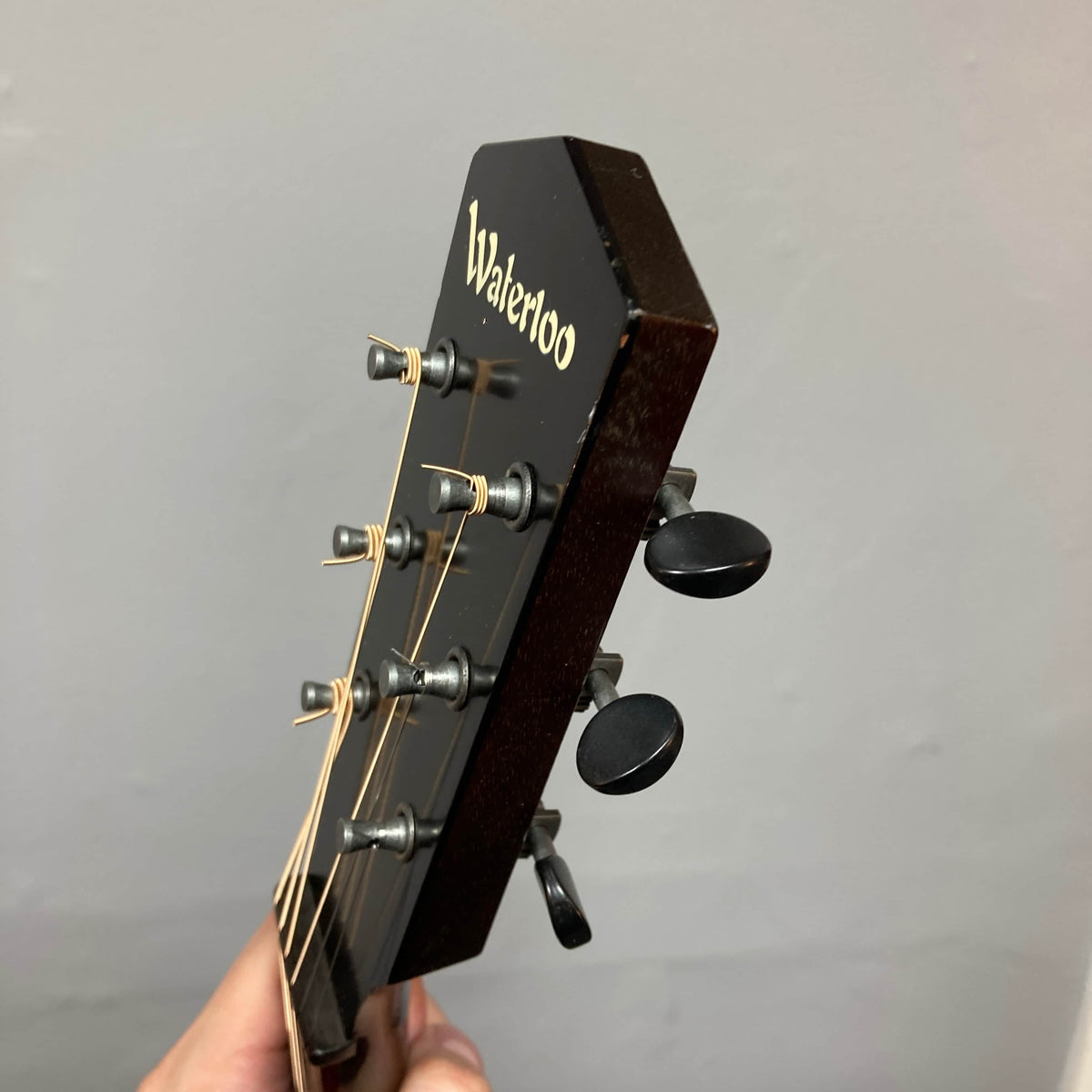 A close-up of the headstock of a Waterloo WL-12 Parlor Acoustic w/Case Used Consign guitar shows tuning pegs, strings, and the "Waterloo" logo against a plain gray background.