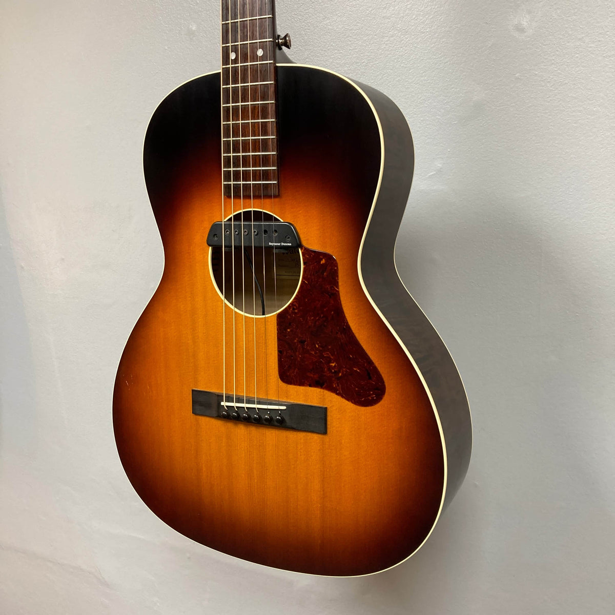 This Waterloo WL-12 Parlor Acoustic w/Case Used Consign features a sunburst finish, six strings, pickguard, and mounted pickup. Photographed against a light wall, it's a vintage-style guitar from Waterloo.