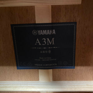 A Yamaha label inside a wooden guitar body displays the model Yamaha A3M ARE Vintage Natural Acoustic Guitar w/Hard Bag Used, indicating it is made in China; it features a solid mahogany build and the advanced SRT2 pickup system.
