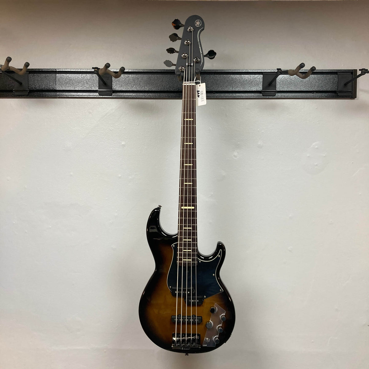 A Yamaha BB735A Electric 5-String Bass in Coffee Sunburst (Blem Discount) by Yamaha hangs on a wall-mounted black guitar rack against a plain light wall, with a tag attached to its headstock.