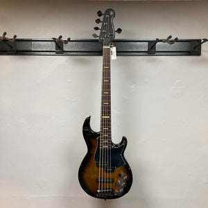 A Yamaha BB735A Electric 5-String Bass in Coffee Sunburst (Blem Discount) by Yamaha hangs on a wall-mounted black guitar rack against a plain light wall, with a tag attached to its headstock.