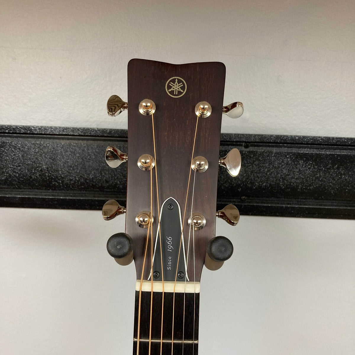 The headstock of a Yamaha FSX5 Satin Red Label Natural acoustic electric guitar, made in Japan with six tuning pegs and a "Since 1966" truss rod cover, is pictured against a white wall in a black guitar holder.
