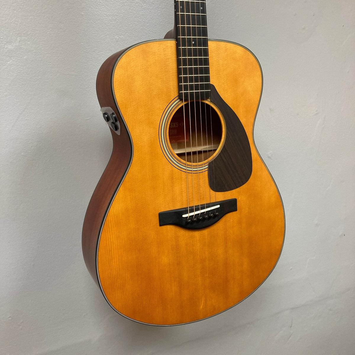 A Yamaha FSX5 Satin Red Label Natural acoustic electric guitar by Yamaha, made in Japan and sold used with case, is shown hanging against a plain light wall. The six-string guitar features a natural finish and dark pickguard.
