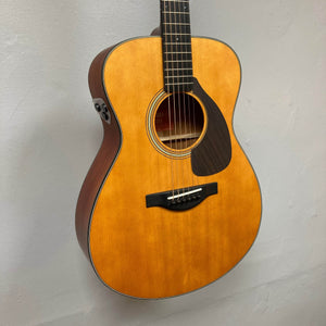 A Yamaha FSX5 Satin Red Label Natural acoustic electric guitar by Yamaha, made in Japan and sold used with case, is shown hanging against a plain light wall. The six-string guitar features a natural finish and dark pickguard.