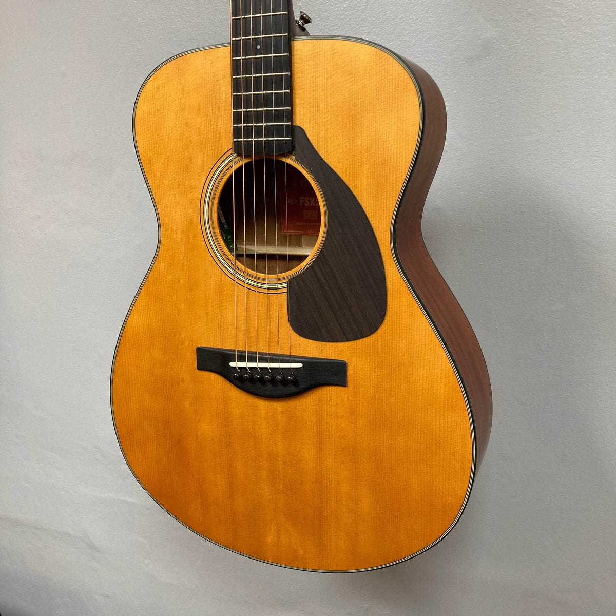 A Yamaha FSX5 Satin Red Label Natural acoustic-electric guitar with a dark pickguard and round sound hole hangs on a light wall. Made in Japan, this guitar features visible steel strings and comes with a case.