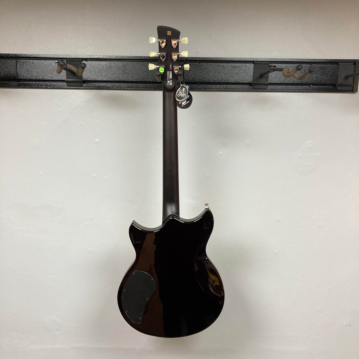 A Yamaha Revstar Standard RSS02T electric guitar in Vintage White, with a chambered mahogany body, hangs on a wall rack. Shown from the back, it has a tag on one tuner and comes with a gigbag.
