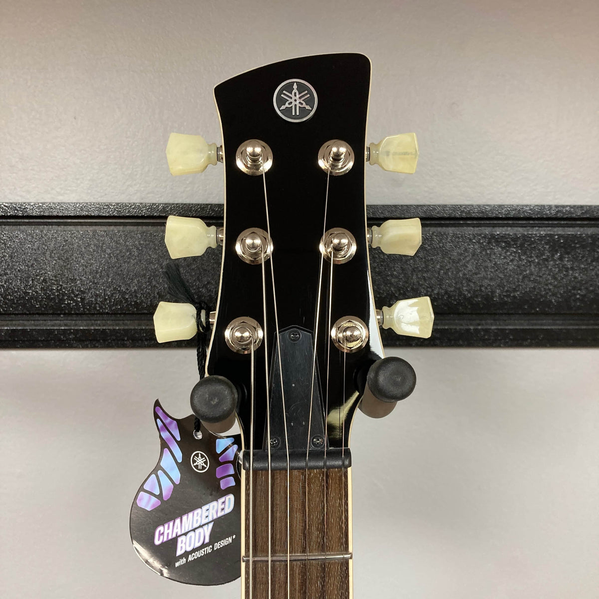 Close-up of the Yamaha Revstar Standard RSS02T Vintage White headstock with six tuners, a "Chambered Body with Acoustic Design" tag, chambered mahogany body detail, and fretboard—all displayed on a wall hanger.
