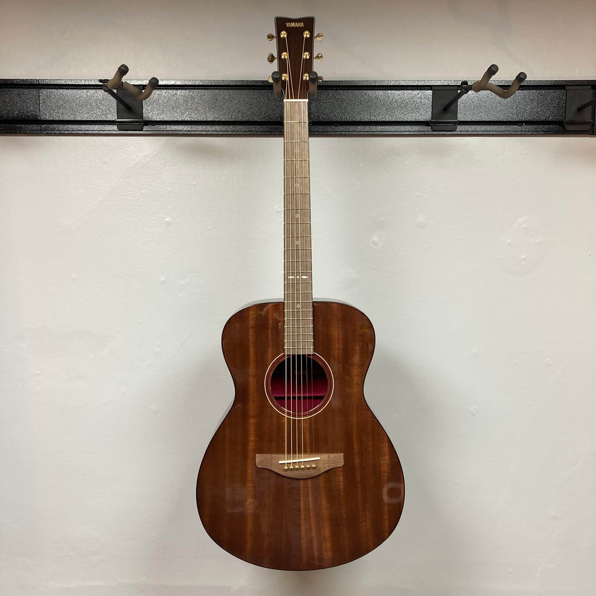 A Yamaha Storia III Solid Mahogany Top Concert guitar hangs on a black wall-mounted holder against a plain white wall.