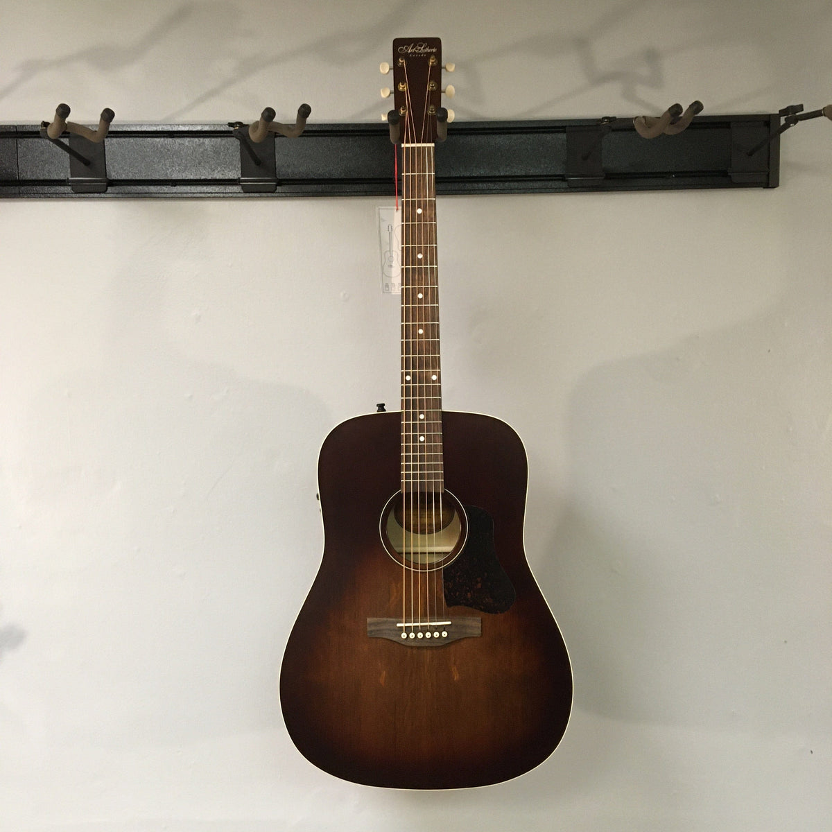 Art & Lutherie Americana Bourbon Burst Acoustic-Electric Guitar on a wall, showcasing its Dreadnought shape and craftsmanship.