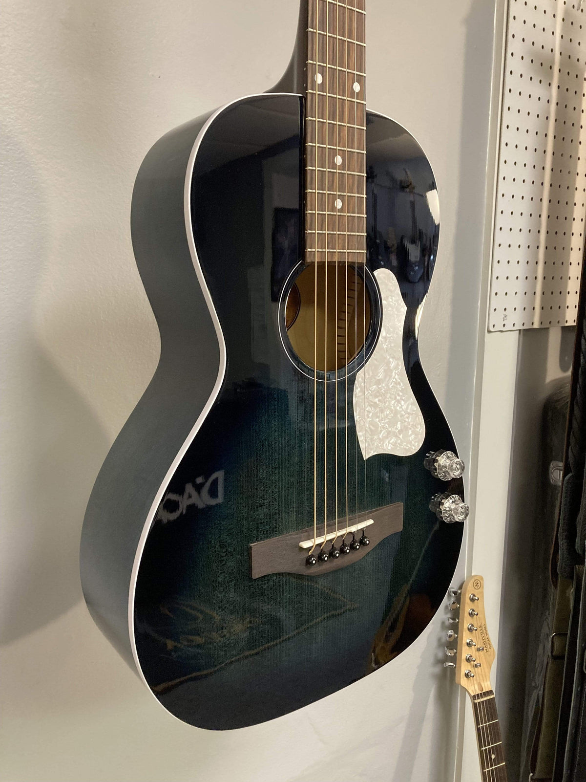 An Art & Lutherie Roadhouse HG Indigo Burst Q-Discrete guitar from Art & Lutherie, featuring a parlor style and solid spruce top, hangs on the wall next to another guitar.