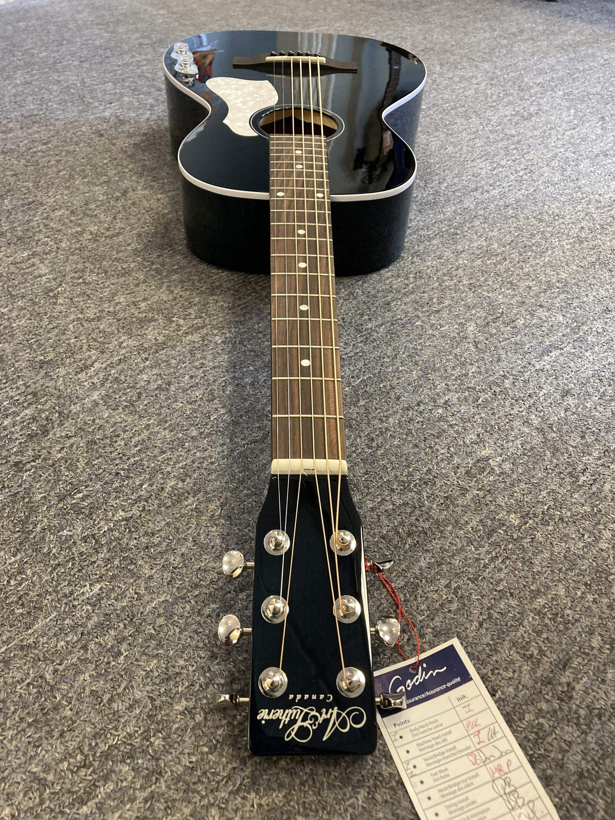 A parlor-style Art & Lutherie Roadhouse HG Indigo Burst Q-Discrete guitar with six strings rests on a gray carpet, featuring an attached tag on the headstock and a solid spruce top for enhanced tone and resonance.