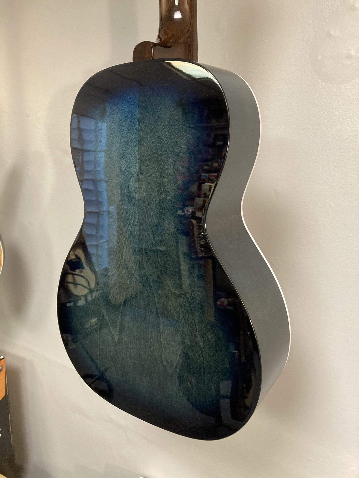 Back of an Art & Lutherie Roadhouse HG Indigo Burst Q-Discrete guitar by Art & Lutherie, parlor-style in dark blue with a glossy finish, reflecting windows and objects, displayed wall-mounted.