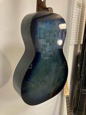 A close-up side view of the Art & Lutherie Roadhouse HG Indigo Burst Q-Discrete, a glossy blue parlor guitar with a solid spruce top by Art & Lutherie, hanging on a wall and reflecting light and nearby objects.