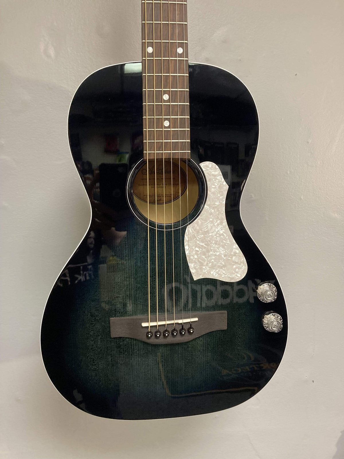 An Art & Lutherie Roadhouse HG Indigo Burst Q-Discrete parlor guitar with two control knobs and a white pickguard hangs on a light-colored wall.
