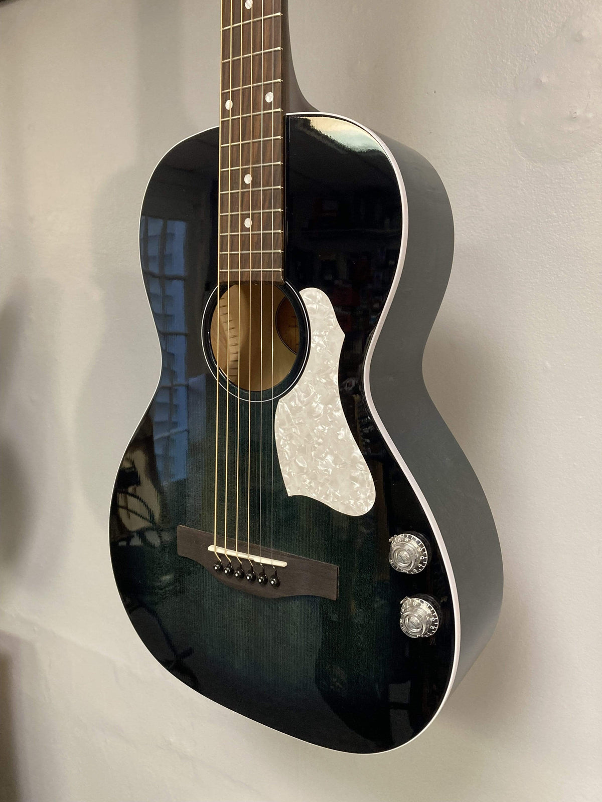 The Art & Lutherie Roadhouse HG Indigo Burst Q-Discrete, a parlor-style acoustic guitar with a white pickguard, two control knobs, and Q-Discrete electronics, is mounted on a light-colored wall.