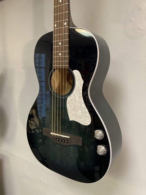 The Art & Lutherie Roadhouse HG Indigo Burst Q-Discrete, a parlor-style acoustic guitar with a white pickguard, two control knobs, and Q-Discrete electronics, is mounted on a light-colored wall.