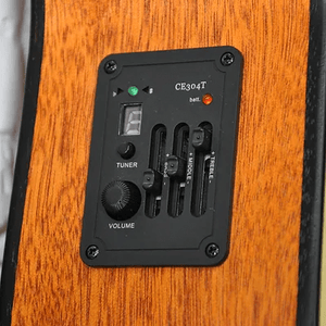 A close-up of the Bodhi BG10E Spruce Top Concert Body Acoustic-Electric Guitar shows its built-in preamp and EQ panel with a tuner display, volume knob, bass/mid/treble sliders, and battery indicator on the wood-finished body.