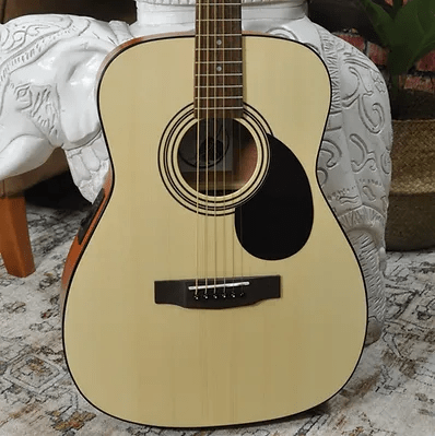 The Bodhi BG10E Spruce Top Concert Body Acoustic-Electric Guitar by Bodhi, with a light spruce top and black pickguard, rests against a white elephant stool on a patterned rug. A woven basket and green plant complete the cozy fingerpicking scene.