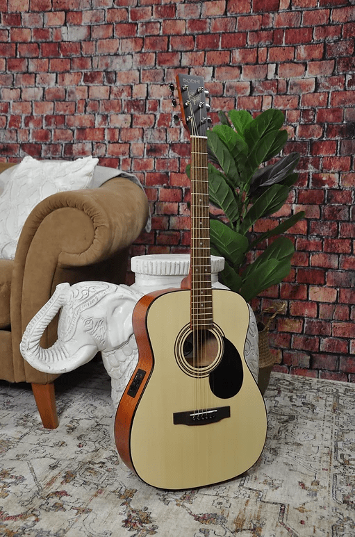 A Bodhi BG10E Spruce Top Concert Body Acoustic-Electric Guitar with a light front and dark sides rests against an elephant-shaped white side table. A tan sofa, green plant, and red brick wall are in the background.