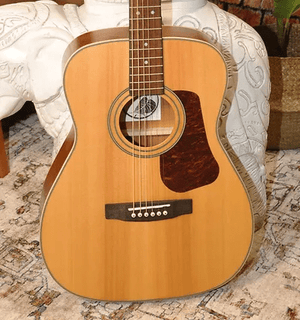 Bodhi BG10S Solid Spruce Top Concert Body Acoustic Guitar on a chair, showcasing its compact design and craftsmanship.