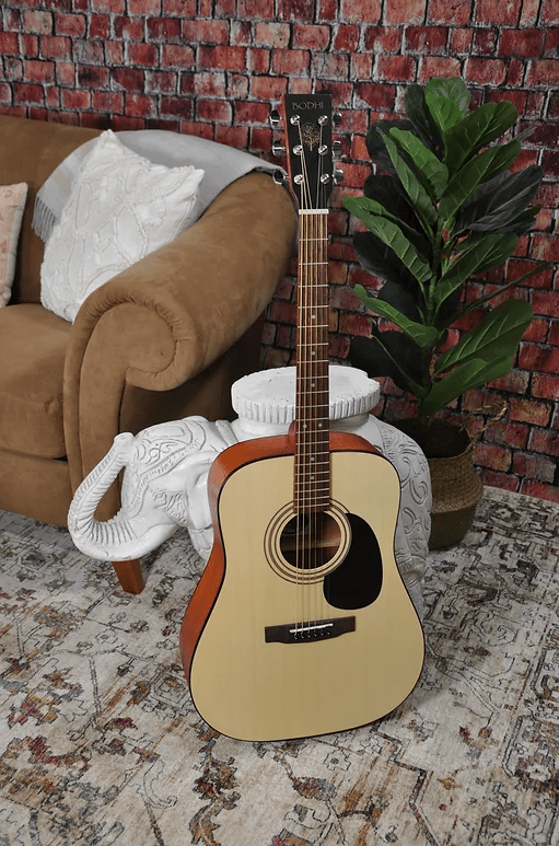 Bodhi BG20 Spruce Top Dreadnaught Acoustic Guitar leaning against a white column, placed on a rug, next to a couch and a potted plant.