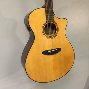 The Breedlove Performer Pro Concerto Aged Toner w/Case, an acoustic guitar by Breedlove with a natural finish and cutaway body known for its dreadnought-like sound, hangs on a light gray wall.